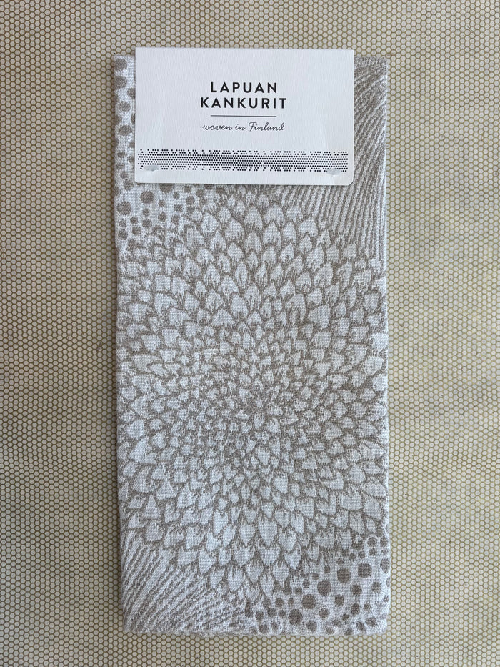 A square woven fabric napkin with a gray and white radial floral pattern, featuring a white paper label at the top that reads 'LAPUAN KANKURIT' and 'woven in Finland'. The napkin is laid flat on a textured beige surface.