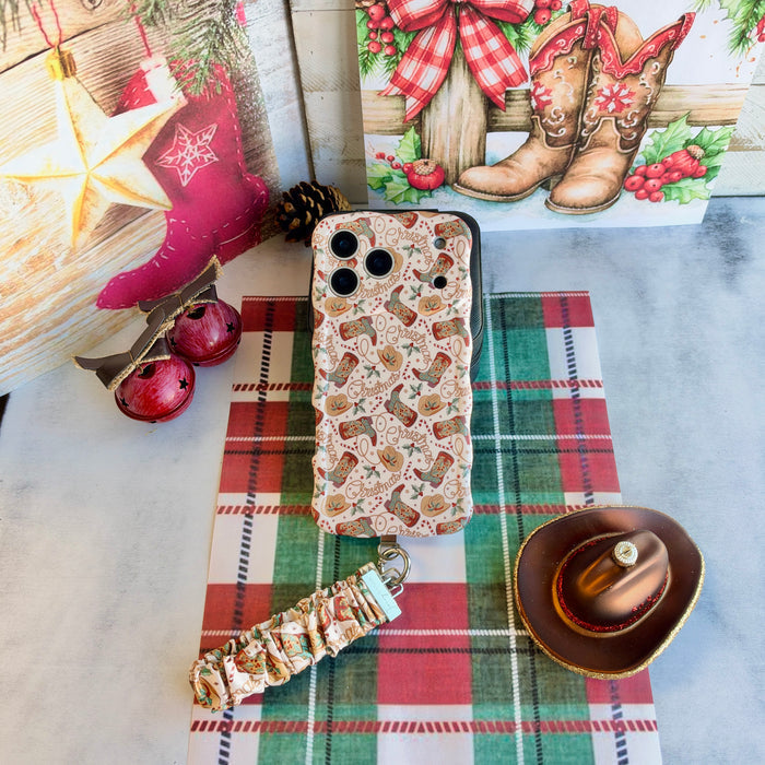 Flat lay of the Boot Scootin' Christmas iPhone case with its cowboy boot scrunchie wrist strap, a pink stocking and cowboy hat ornament