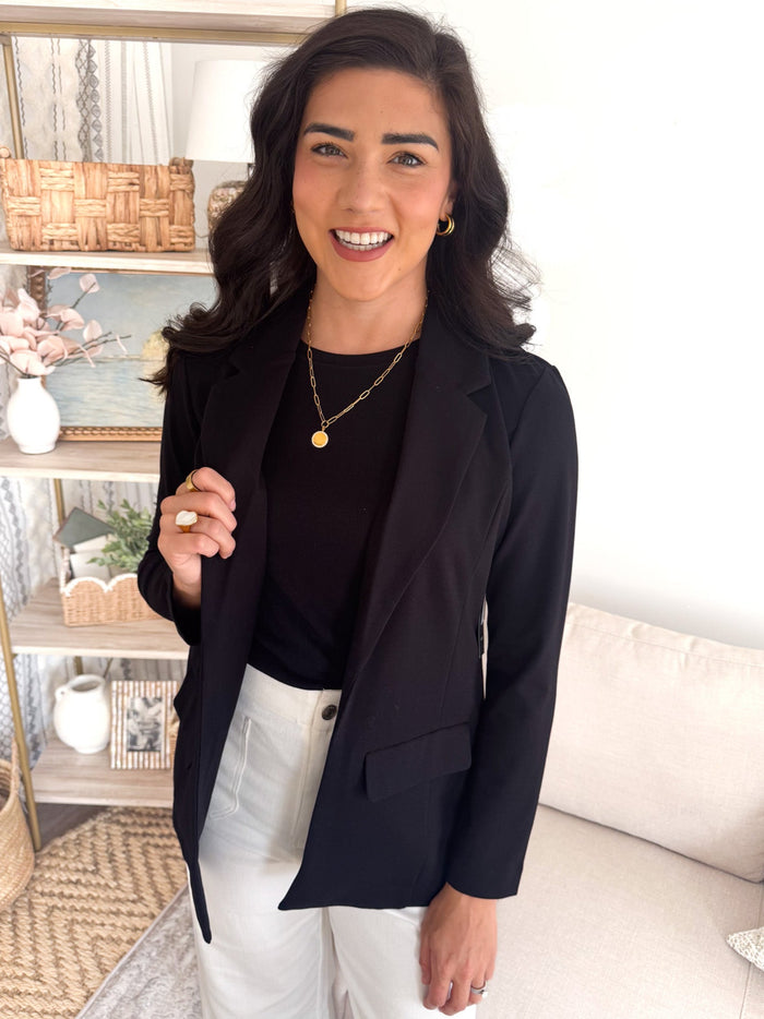 Woman wearing a black blazer over a black top and white pants, standing in a room with a couch and decorative items.