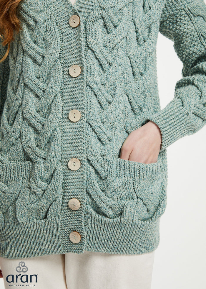 A close-up of a person wearing a sage green cable-knit cardigan sweater with wooden buttons and front pockets, paired with off-white pants. The brand logo 'aran' is visible in the bottom-left corner.