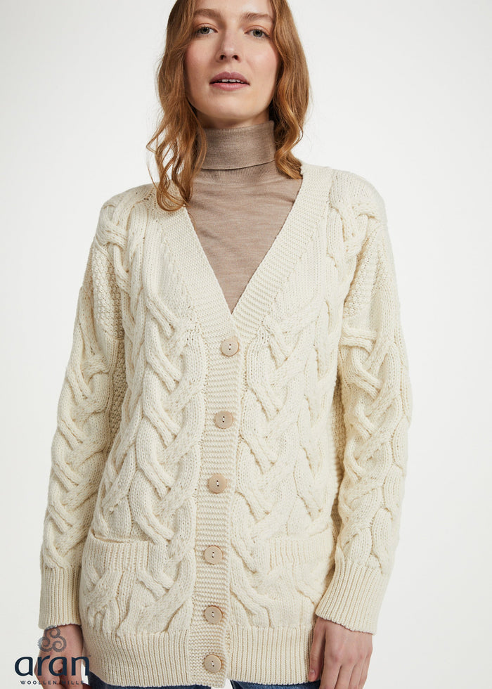 A woman wearing a cream cable-knit cardigan over a beige turtleneck sweater and blue jeans, photographed against a plain light background.