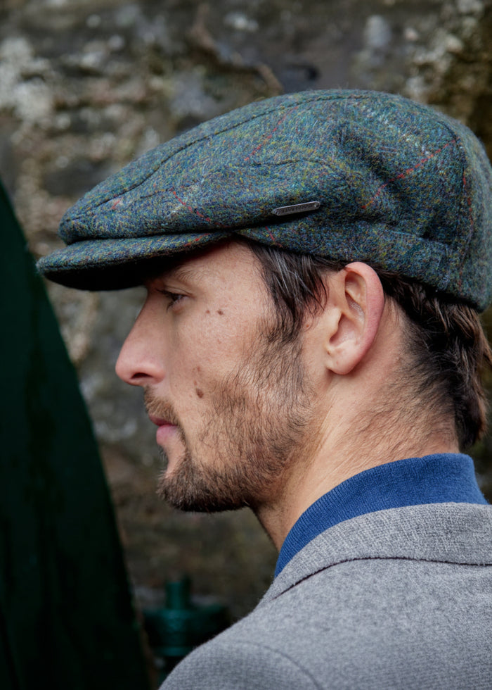 Man wearing a green tweed flat cap against a stone wall background