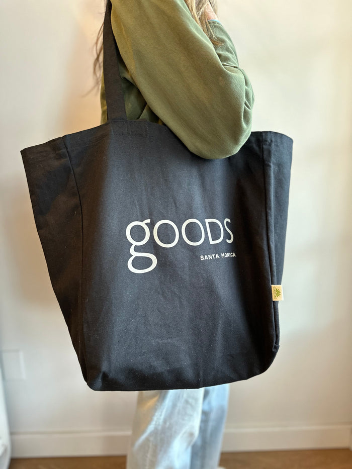 A person holding a black cotton tote bag with the brand name 'GOODS' printed in white on the front.