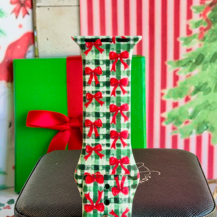 Close-up of a single Plaid Tidings & Bows Apple Watch band strap, red bows on green gingham, in front of a green gift box