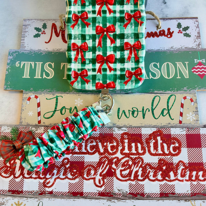 Plaid Tidings & Bows scrunchie phone wrist strap clipped below the matching iPhone case on Tis the Season and Believe in the Magic of Christmas signs