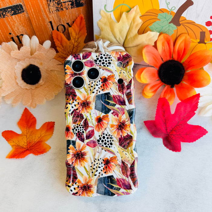 Sienna Petals Wavy Luxe MagSafe iPhone case laid flat among silk sunflowers and autumn maple leaves, showing the sculpted wavy edges and cream floral print.